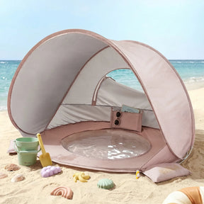The Tiny Land® Baby Beach Tent & Sand Toy Set by Tiny Land—a compact pink and white tent with a built-in mini pool—rests on the sandy shore near the ocean, accompanied by a cheerful sand toy set and seashells, making it the ideal toddler beach haven for seaside fun.|Pink