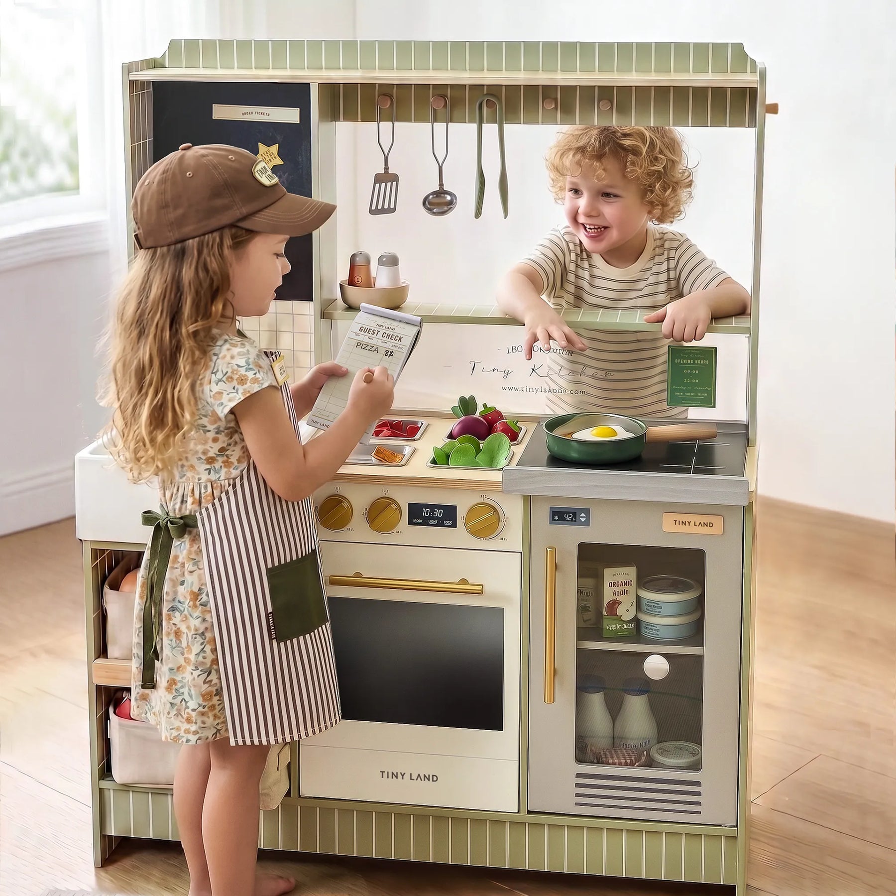 Two children enjoy imaginative play with the Tiny Land® BistroSwitch Play Kitchen. The girl, in a hat and apron, holds a box while the boy in stripes smiles from behind the counter, surrounded by toy food and utensils from Tiny Land.|Green