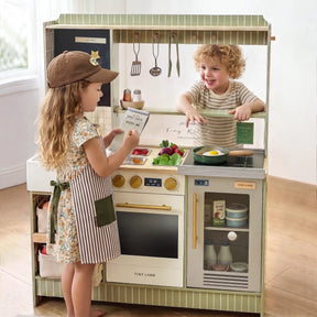 Two children enjoy imaginative play with the Tiny Land® BistroSwitch Play Kitchen. The girl, in a hat and apron, holds a box while the boy in stripes smiles from behind the counter, surrounded by toy food and utensils from Tiny Land.|Green