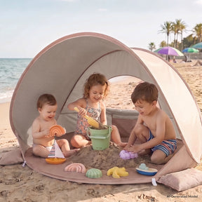 Three children play in the Tiny Land® 3-in-1 Baby Beach Tent, building with colorful sand toys. The beach and ocean are visible nearby as the sun shade from Tiny Land keeps them cool and comfortable while they have fun together.