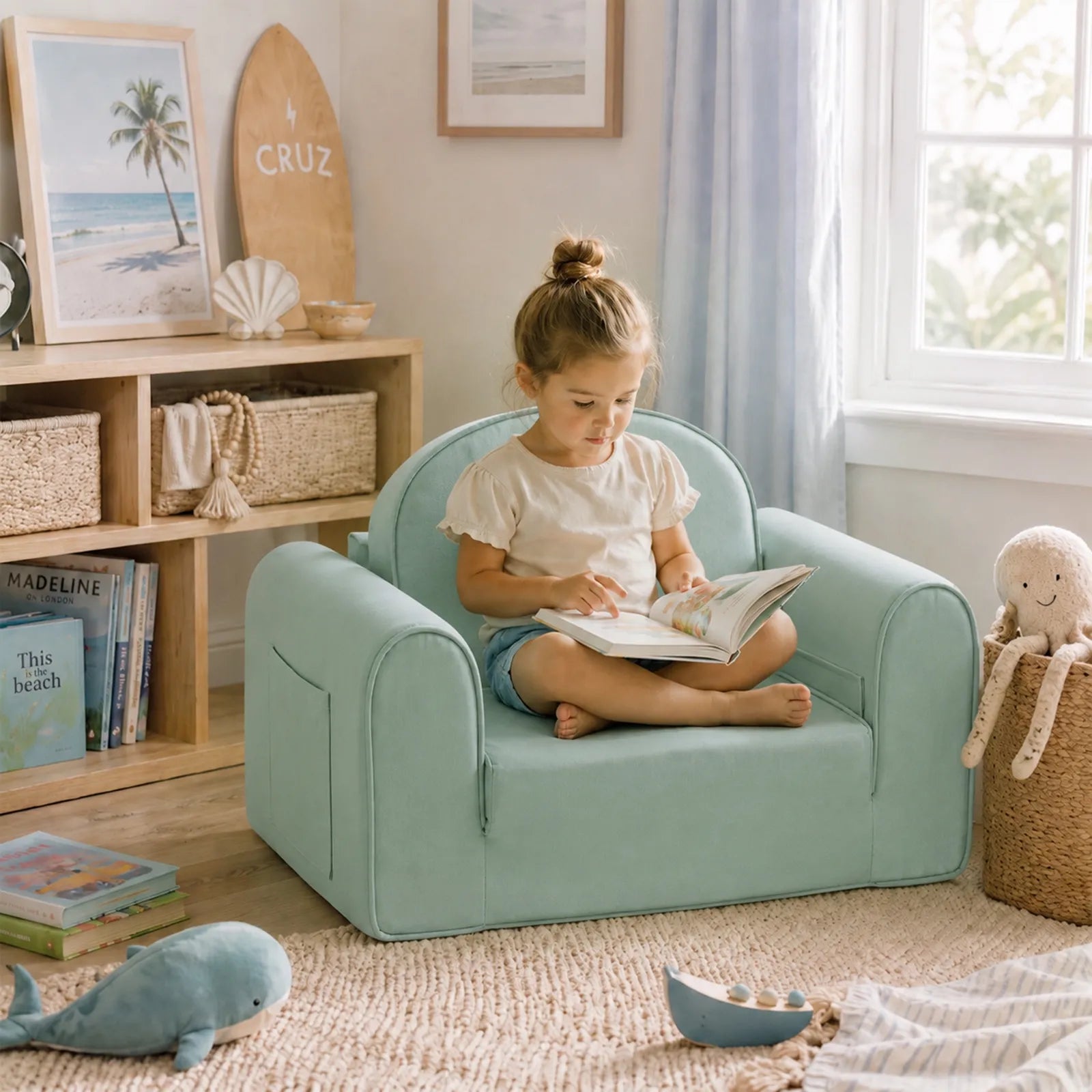 A young girl sits cross-legged on the Tiny Land® GentleSnug Kids Sofa Chair, reading a book in a cozy, sunlit room filled with toys, baskets, books, and a plush octopus by the window.