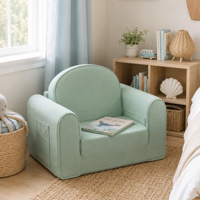 The Tiny Land® GentleSnug Kids Sofa Chair in mint green sits by a sunny window with a book on its seat, near a woven basket, a small decor-filled bookshelf, and a bed with white bedding.