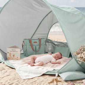 A baby naps on a blanket inside the Tiny Land® Baby Beach Tent by Tiny Land. Close by are a diaper bag, a baby bottle, and a soft book titled "My First Beach Adventure." The included colorful sand toy set from Tiny Land sits ready nearby, promising fun after naptime. This cozy scene is set on sandy beach with the ocean in the background.|Green