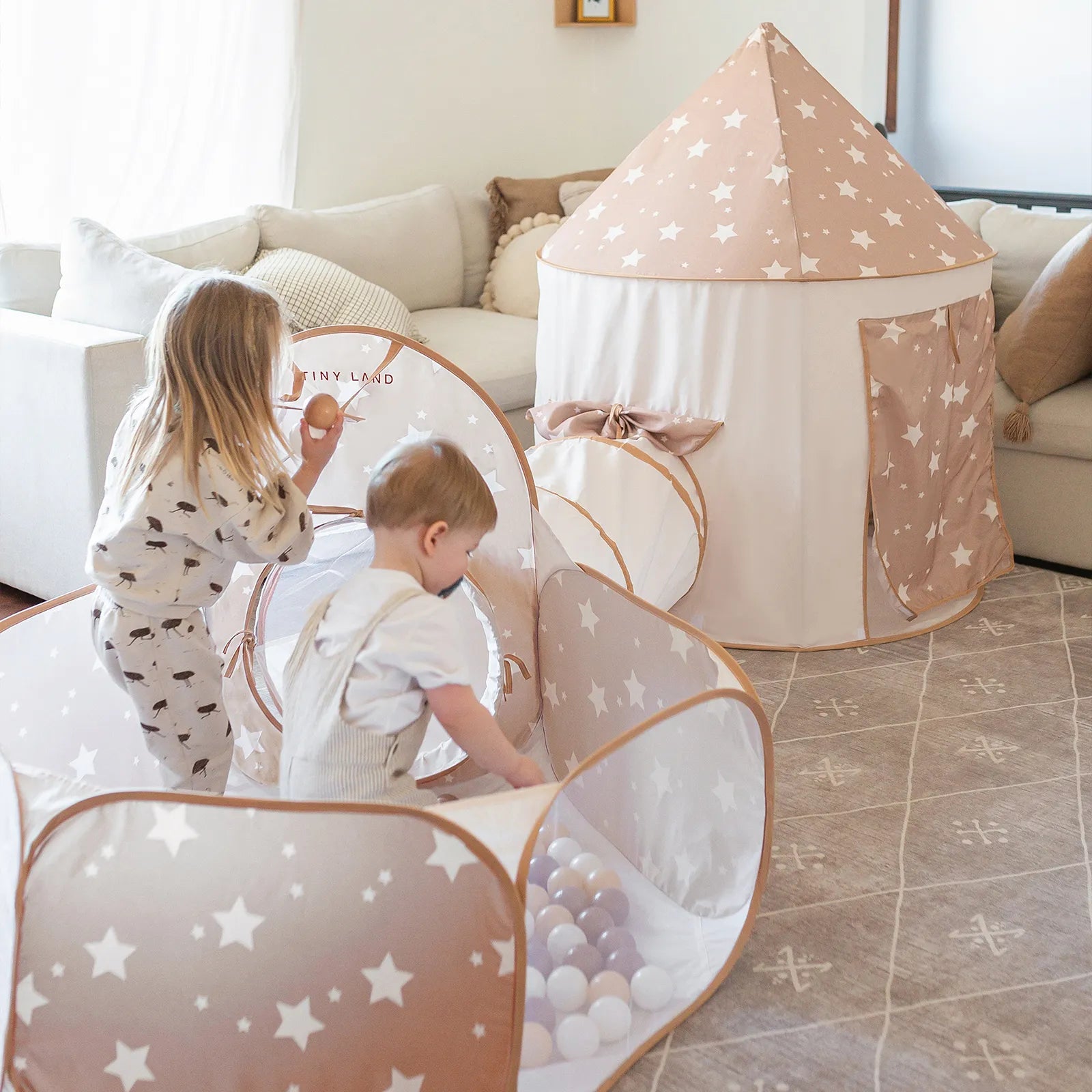 Two young children crawl and play in a star-patterned ball pit inside their playpen, while the Tiny Land® Compact Adventure 3-in-1 Play Tunnel stands nearby in matching beige and white. The cozy, sunlit living room features a comfortable couch and soft rug.|brown