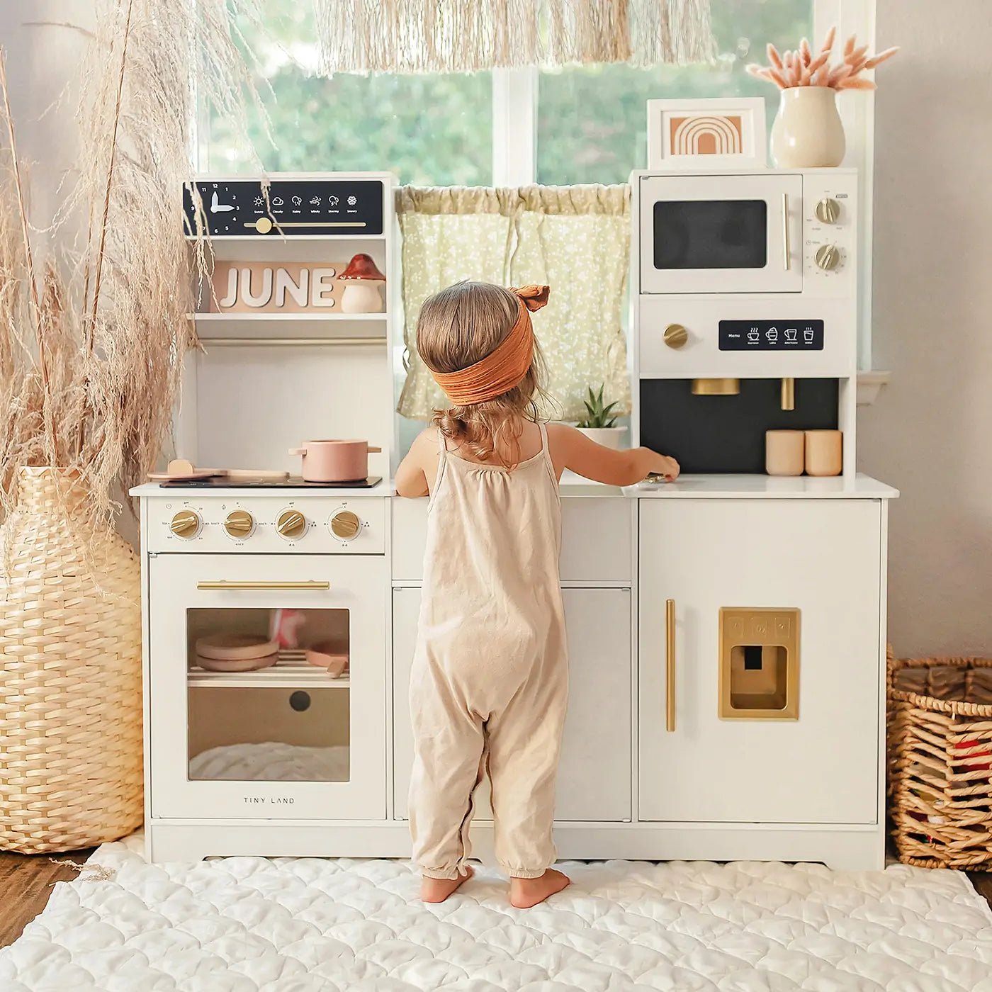 A young child in a peach headband and beige romper reaches for the countertop of the Tiny Land® Iconic Wooden Play Kitchen – Cream Pretend Cooking Set as sunlight streams through a window with a green curtain behind them.