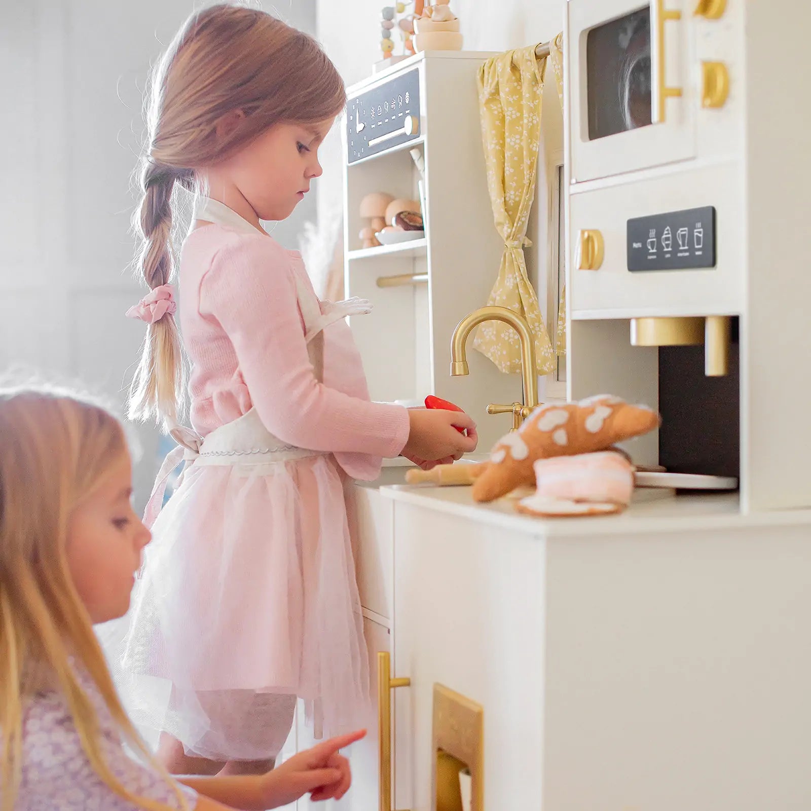 Two young girls play with the Tiny Land® Iconic Wooden Play Kitchen – Cream Pretend Cooking Set for Kids. One stands in a pink dress and apron using a toy utensil, while the other kneels nearby. Toy cookies and pastries are on the counter.