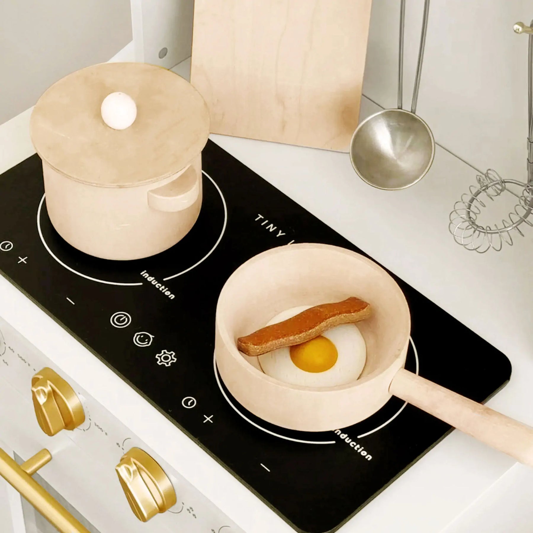 The Tiny Land All-in-One Family Feast Gift Set features a luxury kitchen with black stovetop, wooden pot and pan (with fried egg and bacon), metal utensils, gold cabinet knobs, fridge, and organic food—perfect for kids' cooking fun!.