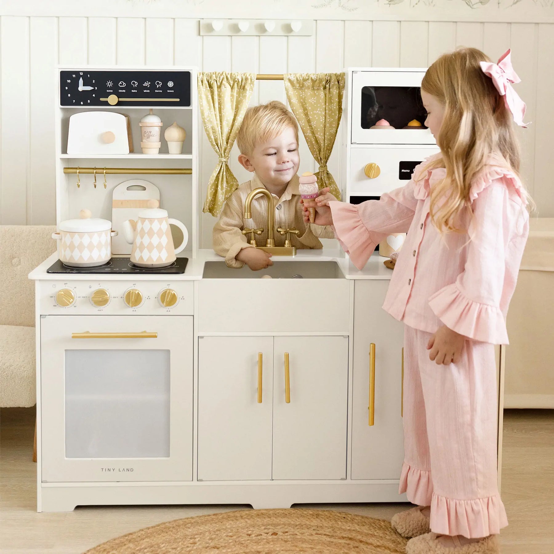 Two children enjoy the Tiny Land All-in-One Family Feast Gift Set; the boy stands behind the kitchen sink, hands in basin, while the girl in pink pajamas pretends to pass him an item near the faucet.