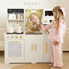 Two children enjoy the Tiny Land All-in-One Family Feast Gift Set; the boy stands behind the kitchen sink, hands in basin, while the girl in pink pajamas pretends to pass him an item near the faucet.