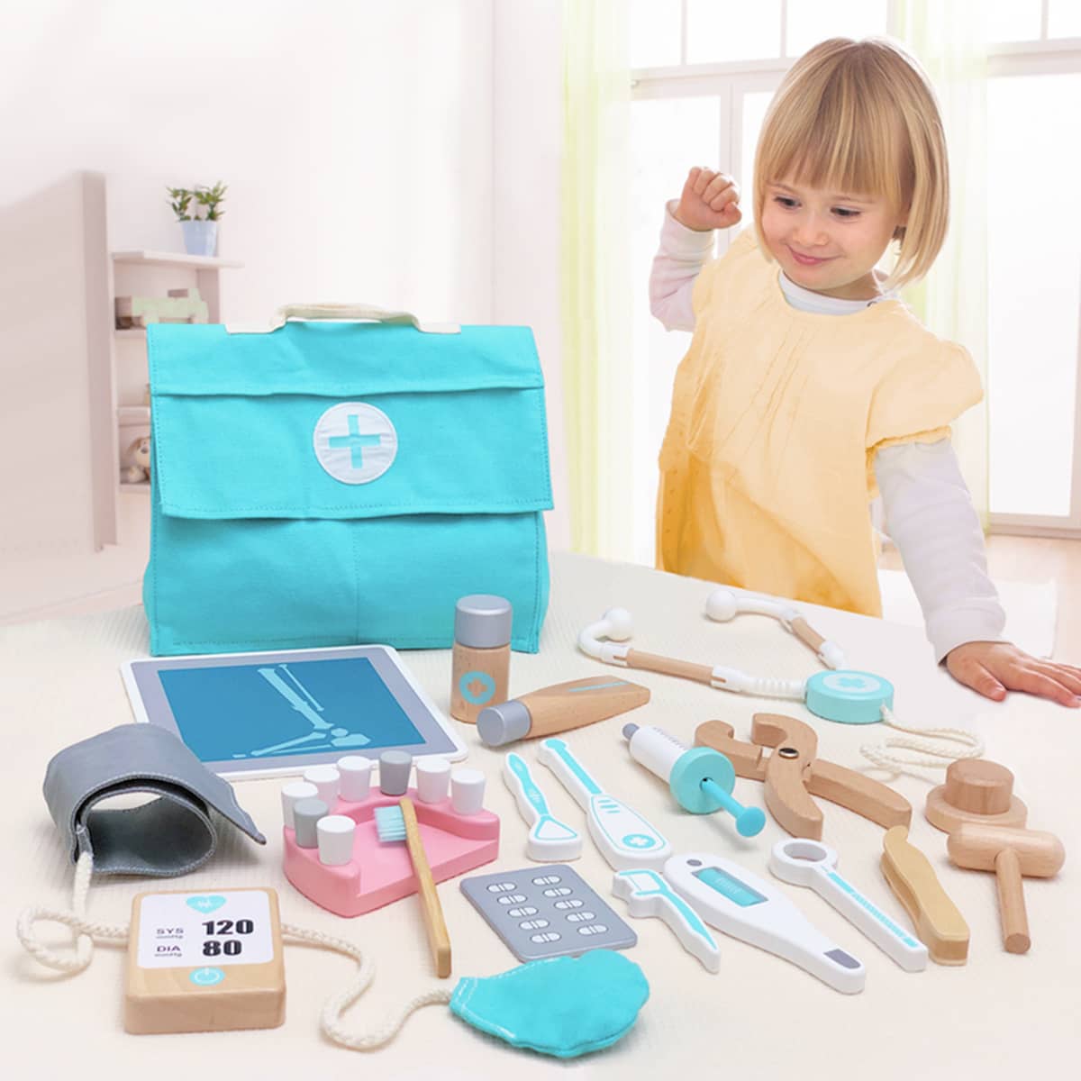 A young child in a yellow smock smiles behind a table with the Tiny Land® Doctor Kit for Kids—by Tiny Land—which features toy medical instruments, a turquoise doctor’s bag, and toy X-ray for imaginative play in a bright room.