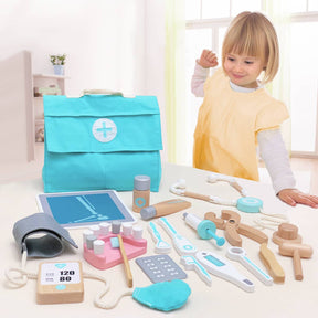 A young child in a yellow smock smiles behind a table with the Tiny Land® Doctor Kit for Kids—by Tiny Land—which features toy medical instruments, a turquoise doctor’s bag, and toy X-ray for imaginative play in a bright room.